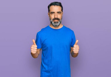Orta yaşlı, sakallı, günlük mavi tişörtlü, başarı işareti taşıyan, elinle olumlu jest yapan, gülümseyen ve mutlu bir adam. Neşeli ifade ve kazanan jest. 