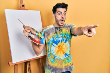 İspanyol asıllı genç adam paletle resim çiziyor. Resim sehpası ayakta duruyor. Parmakları önde, ağzı açık, şaşırmış bir ifade var. 