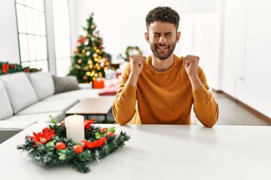 Arap genç adam Noel ağacının yanında masada oturuyor. Başarının heyecanı içinde kolları havada ve gözleri kapalı zaferi kutlarken gülümsüyor. Kazanan konsept. 