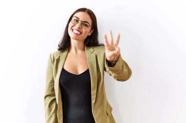 Genç esmer kadın izole edilmiş arka planda duruyor ve üç numaralı parmaklarıyla işaret ederken kendinden emin ve mutlu bir şekilde gülümsüyor.. 