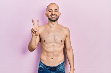 Genç kel adam üstsüz duruyor ve kendini beğenmiş ve mutlu bir şekilde gülümserken iki numarayı işaret ediyor.. 