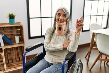 Orta yaşlı, gri saçlı, tekerlekli sandalyede oturan, tasmalı, mutlu gülümseyen, eli ve parmağı yanda olan bir kadın. 