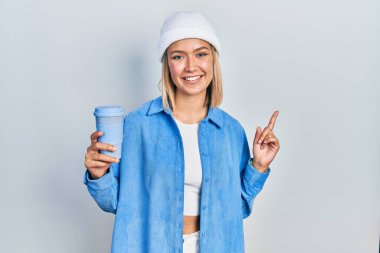 Güzel sarışın kadın bir fincan kahve içiyor gülümsüyor mutlu bir şekilde elini ve parmağını yana doğru gösteriyor. 