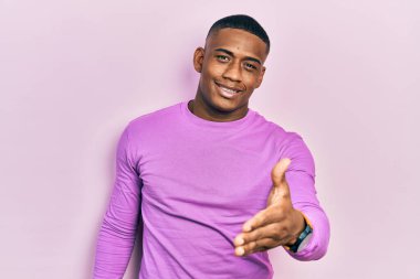 Sıradan pembe kazak giyen genç siyah bir adam dostça gülümseyerek selamlama ve hoş geldin demek için tokalaşıyor. başarılı iş. 