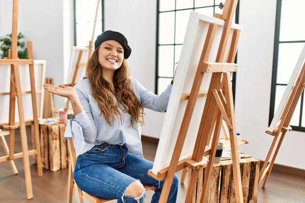 若いヒスパニック系のアーティストの女性は アートスタジオでキャンバスに絵を描く笑顔陽気に提示し カメラを見て手の手のひらで指して — ストック写真