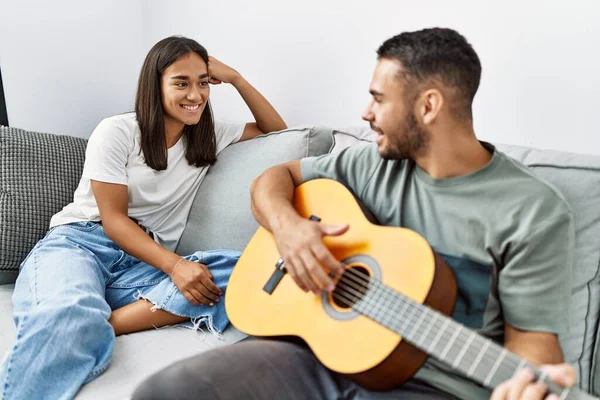 Young latin couple playing classical guitar sitting on the sofa at home.