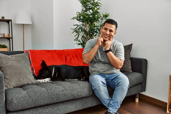 若いですラテン語の男と犬座っていますソファに自宅で笑顔でオープン口 指を指して明るい笑顔を強制 — ストック写真