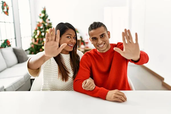 Young Latin Couple Sitting Table Christmas Tree Waiving Saying Hello — Zdjęcie stockowe