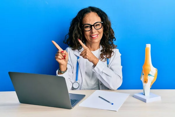 Beautiful middle age woman doctor at orthopedic clinic smiling and looking at the camera pointing with two hands and fingers to the side.