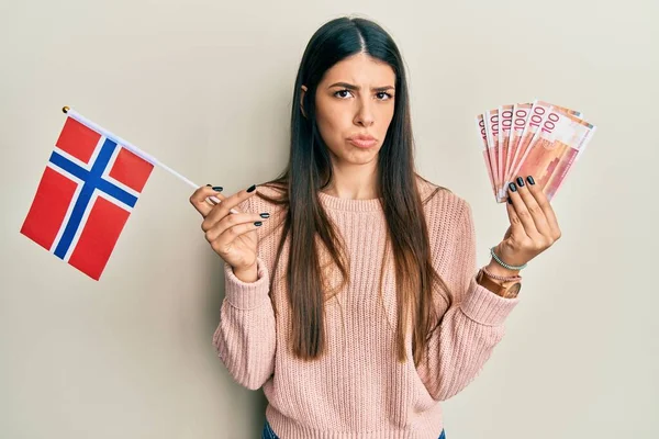 拿着诺威国旗和克朗钞票的年轻惊慌失措的女人表示怀疑和紧张 因为问题皱着眉头 消极的人 — 图库照片