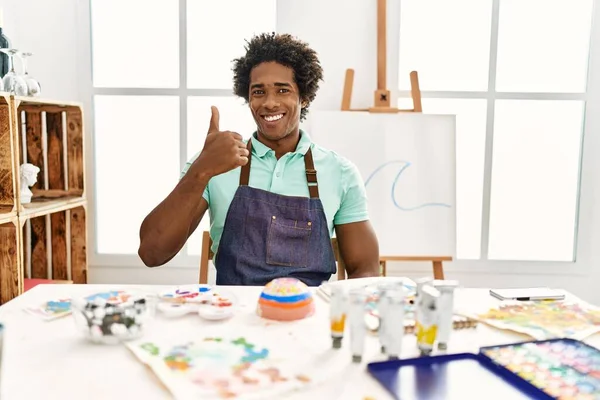 若いアフリカ系アメリカ人の男のテーブルの上にアートスタジオで笑顔幸せと肯定的な 親指アップ行う優れた承認サイン — ストック写真