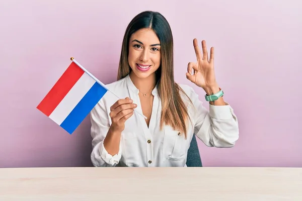 stock image Young hispanic woman holding holland flag sitting on the table doing ok sign with fingers, smiling friendly gesturing excellent symbol 