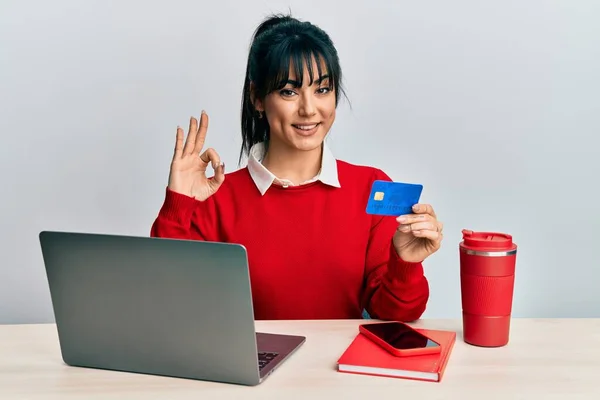 Young brunette woman with bangs working at the office using laptop and credit card doing ok sign with fingers, smiling friendly gesturing excellent symbol