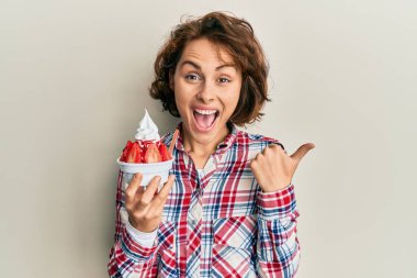 Genç esmer kadın çilekli dondurma yiyor başparmağıyla yan tarafa bakıyor mutlu bir şekilde gülümsüyor. 