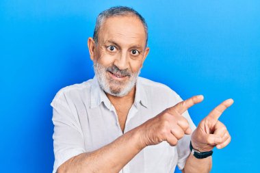 Yakışıklı, sakallı, sıradan beyaz tişörtlü, gülümseyen ve kameraya bakan iki eli ve parmakları yanda olan bir adam.. 