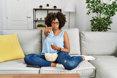 Afro saçlı Afro-Amerikan bir kadın evinde oturmuş patlamış mısır yiyor kamerayı işaret ediyor ve kendine güveni tam bir jest yapıyor. 