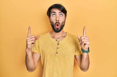 Gündelik giysiler giyen genç İspanyol adam şaşırmış ve şaşırmış bir şekilde yukarı bakıyor ve parmaklarıyla işaret ediyor ve kollarını kaldırıyor.. 