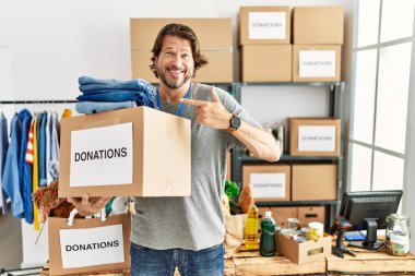 Gönüllü olarak hayır işi için bağış kutusunu tutan yakışıklı orta yaşlı bir adam yüzünde gülümsemeyle mutlu ve doğal bir ifadeyle elini ve parmağını kaldırıyor. 