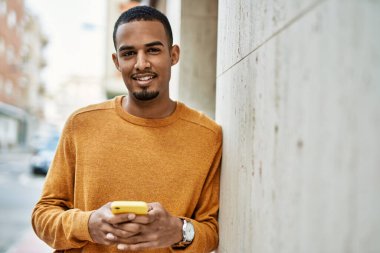 Genç Afrikalı Amerikalı adam şehirde akıllı telefon kullanarak mutlu bir şekilde gülümsüyor..