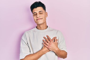 Gündelik beyaz tişört giyen, elleri göğsünde gülümseyen, gözleri kapalı, yüzünde minnet ifadesi olan genç İspanyol bir adam. sağlık kavramı. 