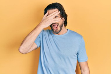 Orta yaşlı beyaz bir adam, günlük kıyafetler giymiş, şok içinde yüzünü ve gözlerini kapatarak utanmış ifadesiyle parmaklarının arasından bakıyor.. 