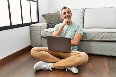 Orta yaşlı İspanyol bir adam oturma odasında dizüstü bilgisayar kullanıyor elleri çenesinde oturmuş soru sormayı, düşünceli ifadeyi düşünüyor. Gülümseyen ve düşünceli bir yüz. şüphe kavramı. 