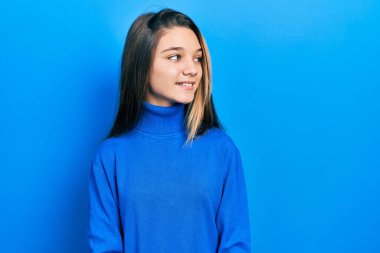 Balıkçı yaka kazak giyen genç esmer kız yüzünde gülümsemeyle yan yana bakıyor, doğal bir ifade. Kendinden emin gülen. 