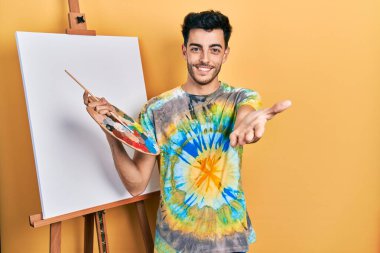 İspanyol asıllı genç adam paletle resim çiziyor. Resim sehpası ayakta gülümsüyor. Avuç içi dolusu yardım ve kabullenme teklif ediyor.. 
