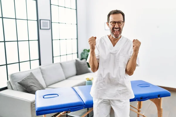 自宅で働いている中年の理学療法士は非常に満足し 腕を上げて勝者のジェスチャーを行う興奮し 笑顔と成功のために叫んでいます お祝いのコンセプト — ストック写真