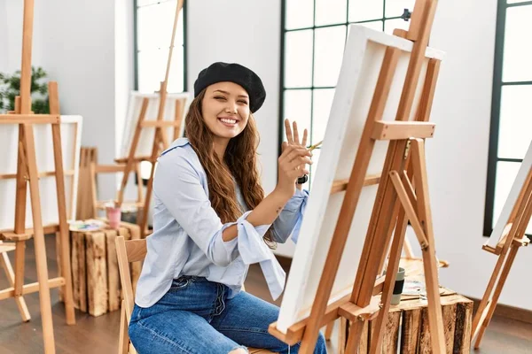 若いヒスパニック系のアーティストの女性は 手と指でOkサインを行う積極的な笑顔アートスタジオでキャンバス上で絵を描く 成功した表現 — ストック写真
