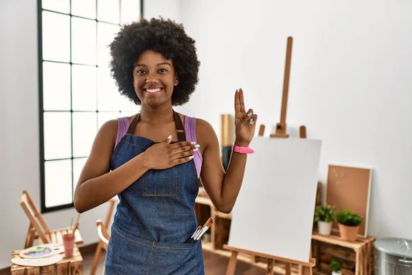 Young african american woman with afro hair at art studio smiling swearing with hand on chest and fingers up, making a loyalty promise oath