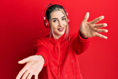 Spor kıyafetleri giymiş, kulaklık takmış, kameraya bakıp kucaklaşmak için gülümseyen genç modern bir kız. Mutluluğu kucaklayan neşeli bir ifade.. 