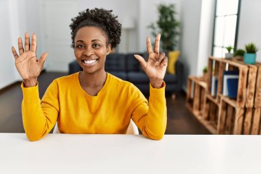 Genç Afro-Amerikalı kadın, gündelik giysiler giymiş evde masanın üzerinde oturuyor ve 8 numarayı gösterirken kendinden emin ve mutlu bir şekilde gülümsüyor.. 