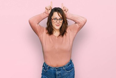 Genç ve cüsseli bir kadın günlük kıyafetler ve gözlük takıyor ve parmağıyla boynuz gibi komik hareketler yapıyor. 