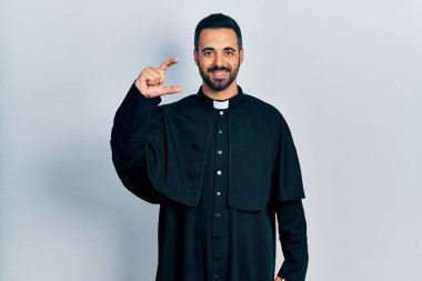 Sakallı yakışıklı, Katolik rahip cüppeli, gülümseyen, kendine güvenen, el hareketi yapan, parmakları küçük gösteren ve kameraya bakan bir adam. ölçüm kavramı. 