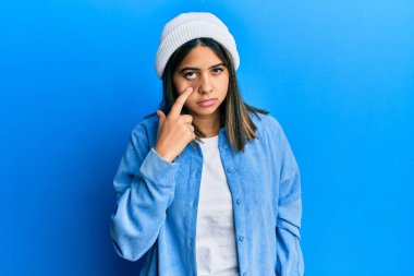 Tatlı yün şapkalı genç Latin kadın senin jestini izlerken şüpheli bir ifade takınıyor. 