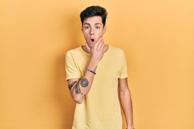 Gündelik sarı tişört giyen genç İspanyol adam inanamıyor, şaşırmış ve elleri çenesinde şaşkın bir ifade takınıyor. 