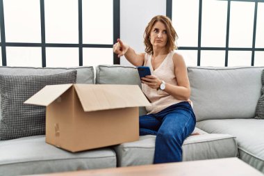 Orta yaşlı bir kadın koltukta oturmuş, teslimat paketini kontrol ediyor, kamerayı işaret ediyor ve sen kendinden emin bir şekilde ciddi görünüyorsun. 