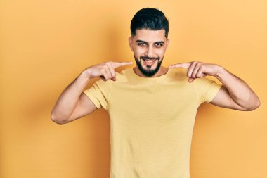 Sakallı, sıradan sarı tişörtlü, neşeli gülümseyen ve parmaklarıyla ve ağzıyla işaret eden yakışıklı bir adam. diş sağlığı kavramı. 