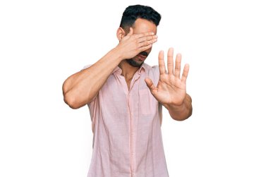 Sakallı, sıradan tişörtlü, elleriyle gözlerini kapatan, üzgün ve korku ifadeli İspanyol bir adam. utangaç ve olumsuz kavram. 