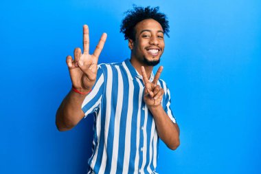 Sakallı, sıradan çizgili tişörtlü, kameraya bakıp zafer işareti yapan Afro-Amerikalı genç bir adam. İki numara.. 