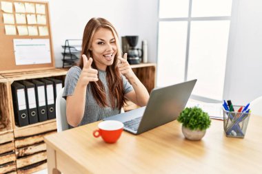 Ofiste çalışan genç esmer kadın dizüstü bilgisayarla kamerayı mutlu ve komik yüzüyle işaret ediyor. iyi enerji ve titreşimler. 