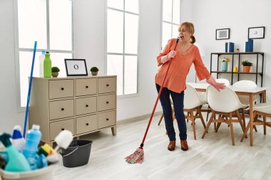 Orta yaşlı temizlikçi kadın gülümseyerek evde paspas kullanarak dans edip şarkı söylüyor..