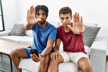 Genç ispanyol adamlar evdeki kanepede otururken akıllı telefon kullanıyorlar ve avuç içiyle şarkı söylemeyi bırakıyorlar. Yüzünde olumsuz ve ciddi bir jest olan uyarı ifadesi. 