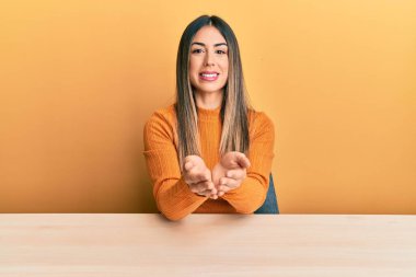 Elleri avuçlarında gülümseyerek masada oturan ya da el hareketi yapan Latin asıllı genç bir kadın. Bekleme ve koruma 
