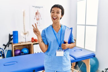 Kısa saçlı, İspanyol bir kadın ağrı kesici kliniğinde çalışıyor. Gülümsüyor, kameraya göz kırpıyor ve parmaklarıyla zafer işareti yapıyor. İki numara.. 