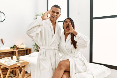 Young latin couple wearing towel standing at beauty center shouting and screaming loud to side with hand on mouth. communication concept. 