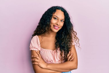 Gündelik kıyafetler giyen, kollarını kavuşturup kameraya gülümseyen genç Latin kadın. pozitif kişi. 