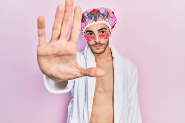 Bornoz ve duş başlığı takan genç İspanyol adam. Göz bantları kullanıyor. El açık yamalar takıyor. 