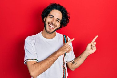 Yakışıklı, beyaz tişört giyen, gülümseyen ve kameraya bakan, iki elini ve parmaklarını yana doğru gösteren İspanyol bir adam.. 